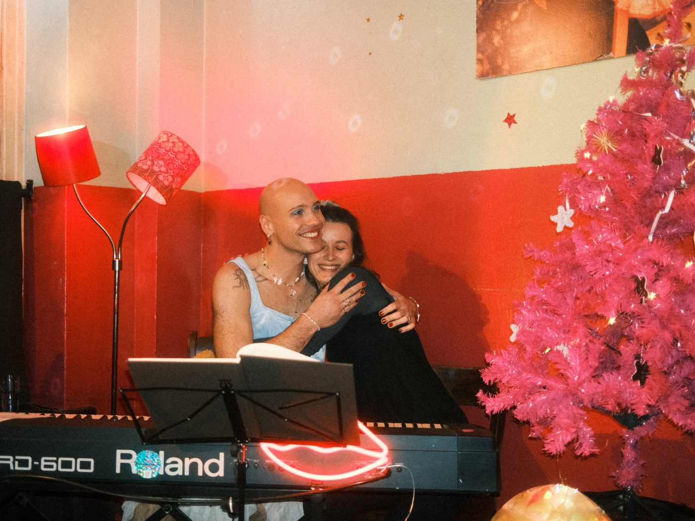 Paula Kahre und Gesa Penthin umarmen sich lachend hinter einem Keyboard mit Notenst&auml;nder, vor einer roten Wand mit Stehlampe, daneben ein pinker Weihnachtsbaum