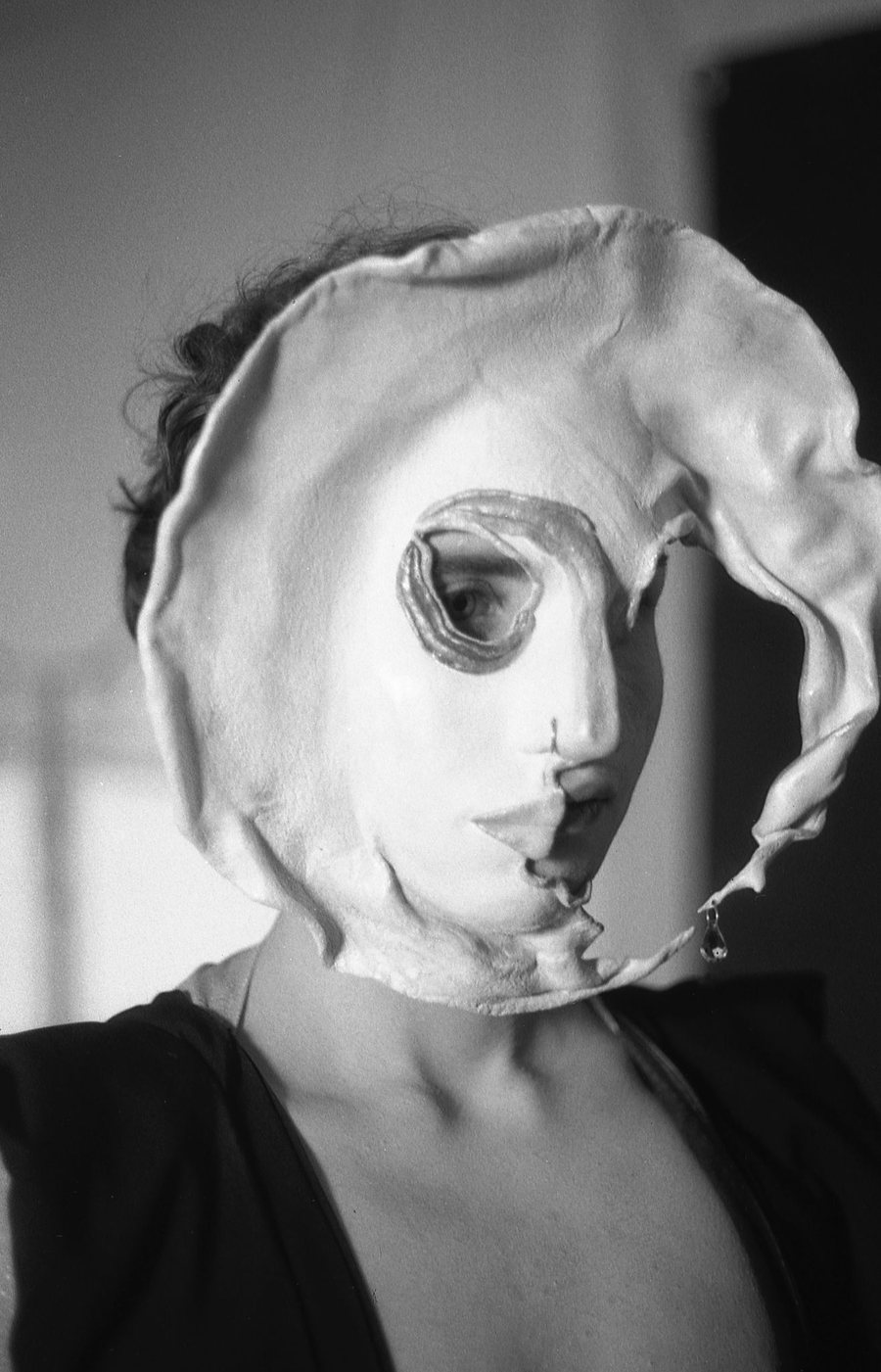Schwarzwei&szlig;-Foto: Person mit einer gro&szlig;en, verformten Maske, durch deren Augenloch ein Auge blickt