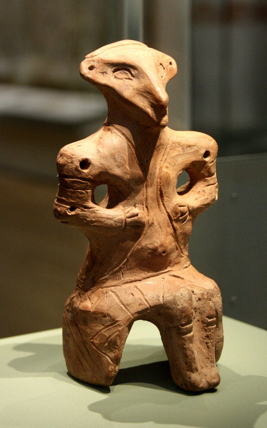 Sitzende Tonfigur mit maskenhaftem, vogelartigem Kopf und gro&szlig;en Augen, der K&ouml;rper mit eingeritzten Linien verziert